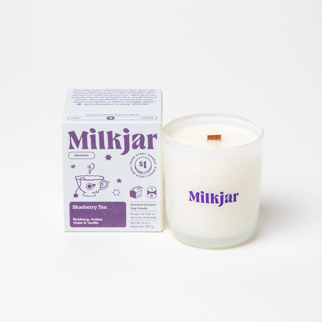 Milkjar candle in a white container placed beside purple packaging with branding on a white background.