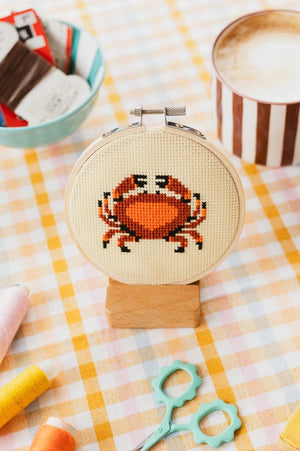 Cross-stitched red and orange crab design on a embroidery hoop with a cup of coffee and snacks in the background.