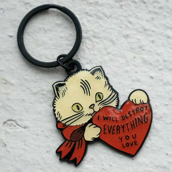 black keychain of a white cat wearing a red bow holding a large red heart. text in the heart reads "i will destroy everything you love"