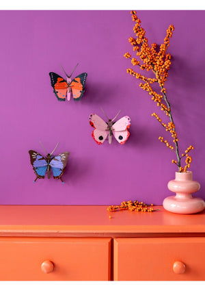 Three pop-up 3D butterfly models hanging on a purple wall with an orange dresser and pink vase.
