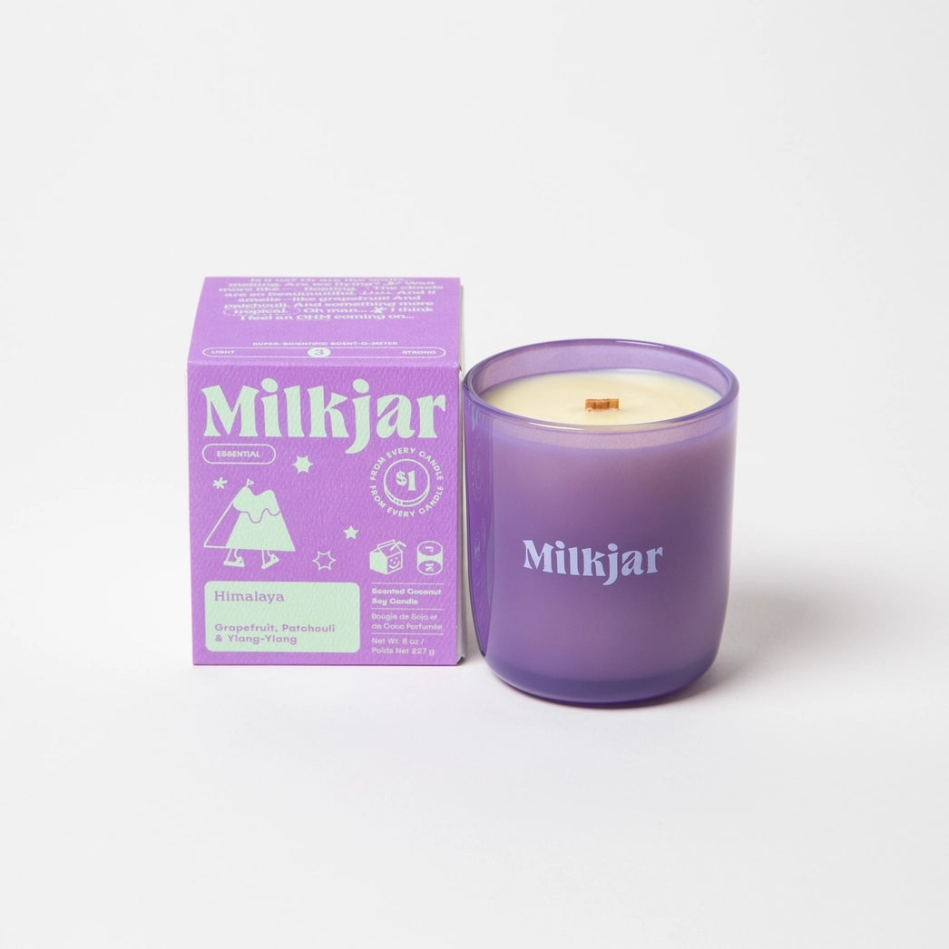 Milkjar candle in a purple container placed beside purple packaging with green branding on a white background.