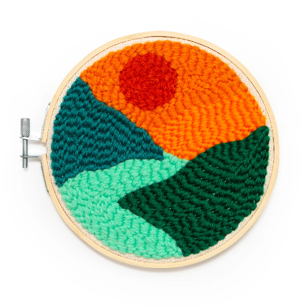bright, simple punch needle landscape in a wooden circle. a red sun is above a turquoise river cutting through hills. kikkerland product on white background