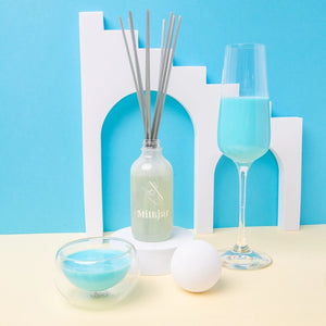 Decorative items including a Milk Jar reed diffuser, a champagne glass, and blue candle in a glass container against a blue background