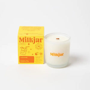 Canadian company Milkjar candle and its box packaging. The white candle is in a translucid glass with "Milkjar" written in yellow. The other side of the candle has a cute drink doodle in yellow. The packaging is a small bright yellow box with orange text and graphics. 