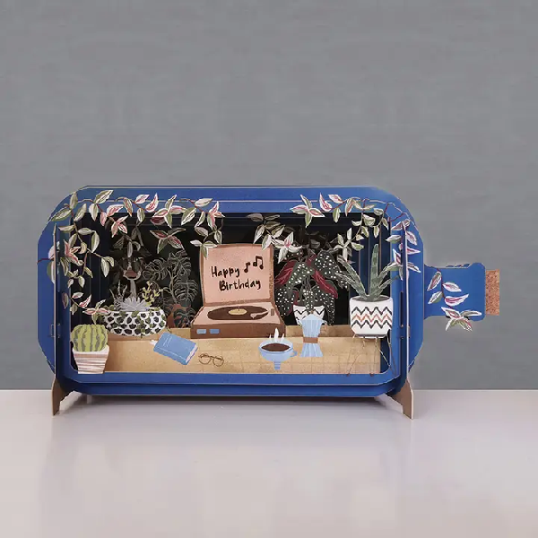 blue bottle pop up card. the intricate paper cuttings depict a homey scene with plants, books, and an open record player with the text "happy birthday"