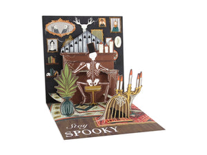 A 3D pop-up cardboard Halloween greeting card on a white background. The card features a skeleton sitting in front of a piano, with a birdcage holding candles, a potted plant, and the text 'Stay SPOOKY' printed on the base.