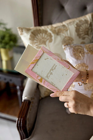 Hand holding a photo frame cotton card with a pink and gold marbled design in a home setting. The text 'Happy Birthday Lovely Person' is printed on the card. 