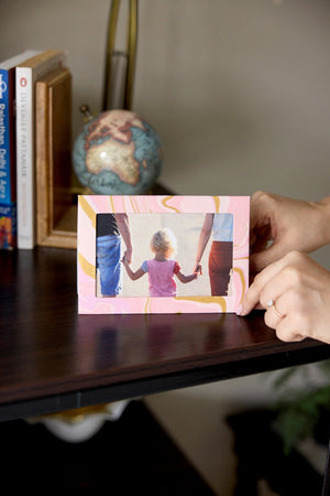 Hand holding a small rectangular photo frame card with a pink and gold marbled design. A photo showing a beach scene is inside the frame. It sits on a wooden surface with books and globes in the background.