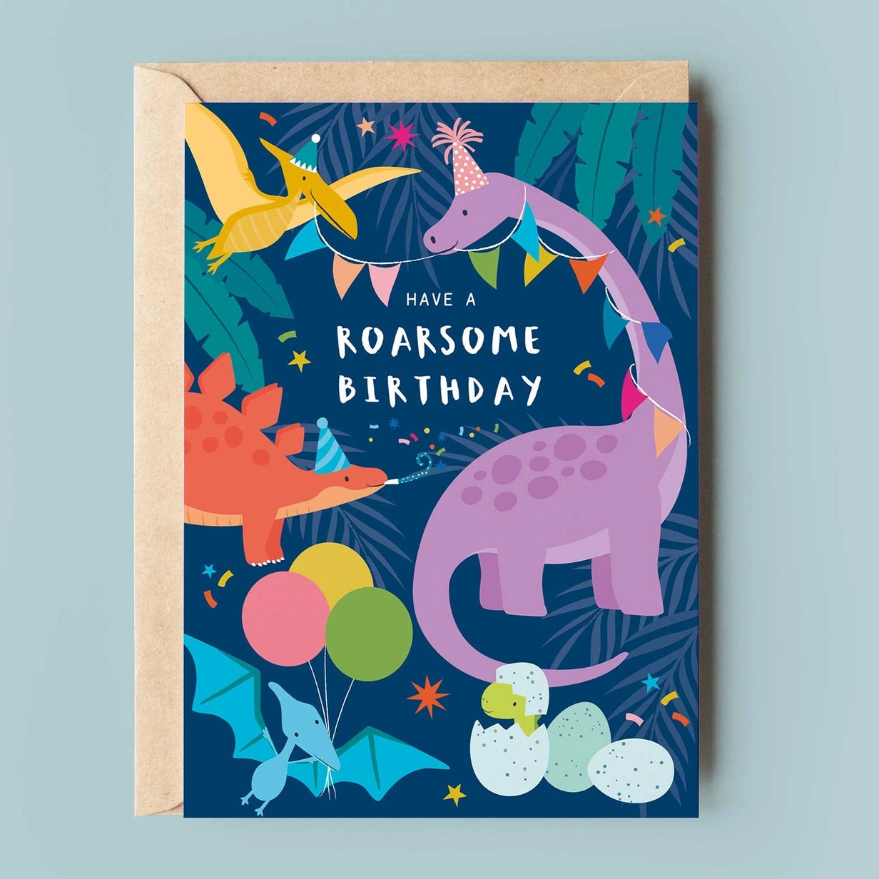 Greeting card featuring colourful illustrations of dinosaurs wearing party hats. Two of the dinosaurs are carrying a garland in their mouths, one holds balloons tied with string, another is blowing a party blower, while a baby dino is hatching from an egg. The text 'Have a roarsome birthday' is printed on a dark blue background with leaf designs.