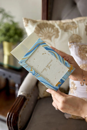 Hand holding a photo frame cotton card with a blue and gold marbled design in a home setting. The text 'Happy Birthday To You' is printed on the card. 