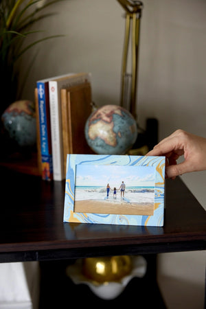 Hand holding a small rectangular photo frame card with a blue and gold marbled design. A photo showing a beach scene is inside the frame. It sits on a wooden surface with books and globes in the background.