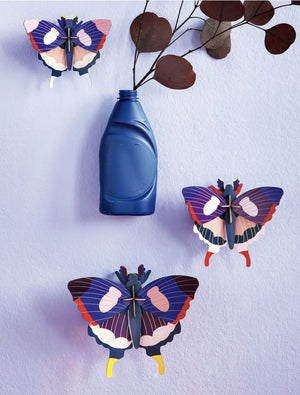 Three 3D butterflies hanging on a light blue background, alongside a bottle containing a plant. The butterflies are blue, pink, white, purple and red in colour. 