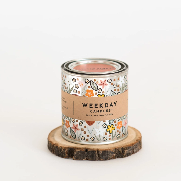 silver tin candle with painted doodles white, brown, peach, and orange flowers. a brown tag wraps the tin. on it, "Weekday Candles 100% Soy Wax Candle", the name of the product, the ingredients and instructions are written in a sharp black font.