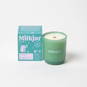 Canadian company Milkjar candle and its box packaging. The white candle is in a green glass with "Milkjar" written in baby blue. The other side of the candle has a cute tree stump doodle in baby blue. The packaging is a small blue box with lilac text and graphics. 