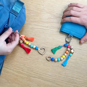 Two colorful keychains with beads and tassels attached to a blue bag and a purse on a wooden surface.