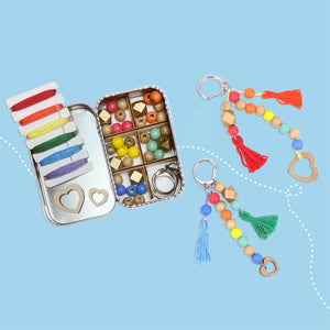 Open container filled with coloured beads and threads, beside two beaded keychains on a blue background. 