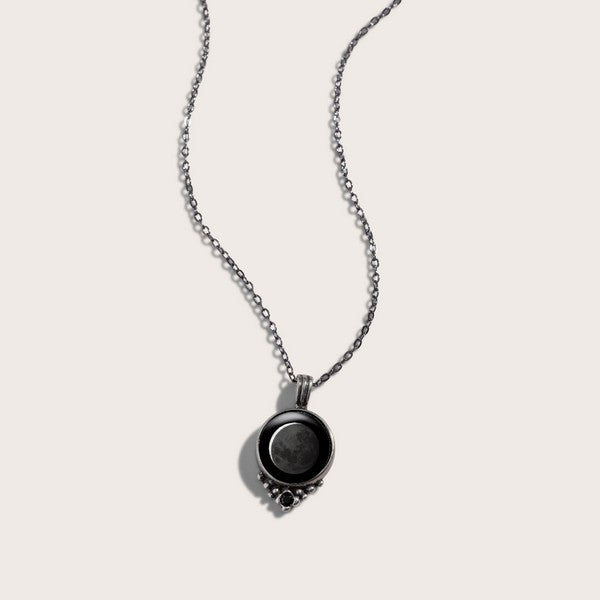 black circle pendant connected to a delicate silver chain. the pendant has an image of the moon in it as well as silver details forming a V at the bottom