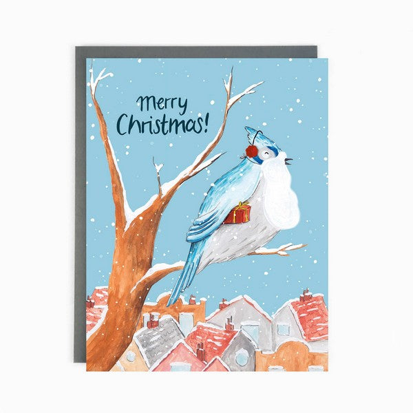 A Christmas card featuring a blue jay perched on a branch with snow falling and a town in the background. The card has the text 'Merry Christmas!' at the top. The blue jay has a gift tucked under it's wing and is wearing a pair of red ear muffs. 